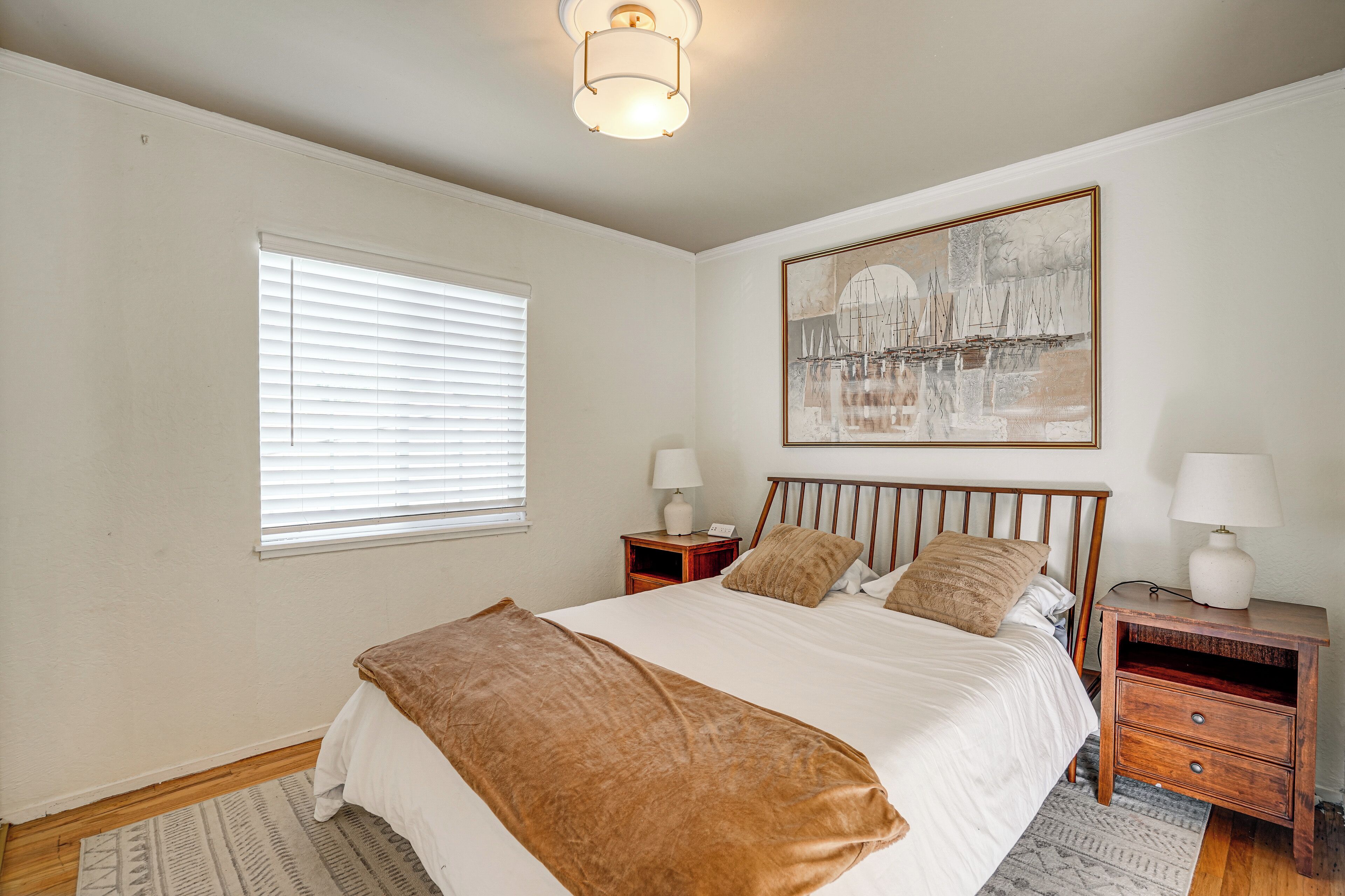 A bedroom with a bed, a nightstand, a lamp, a painting on the wall, and a window with blinds.