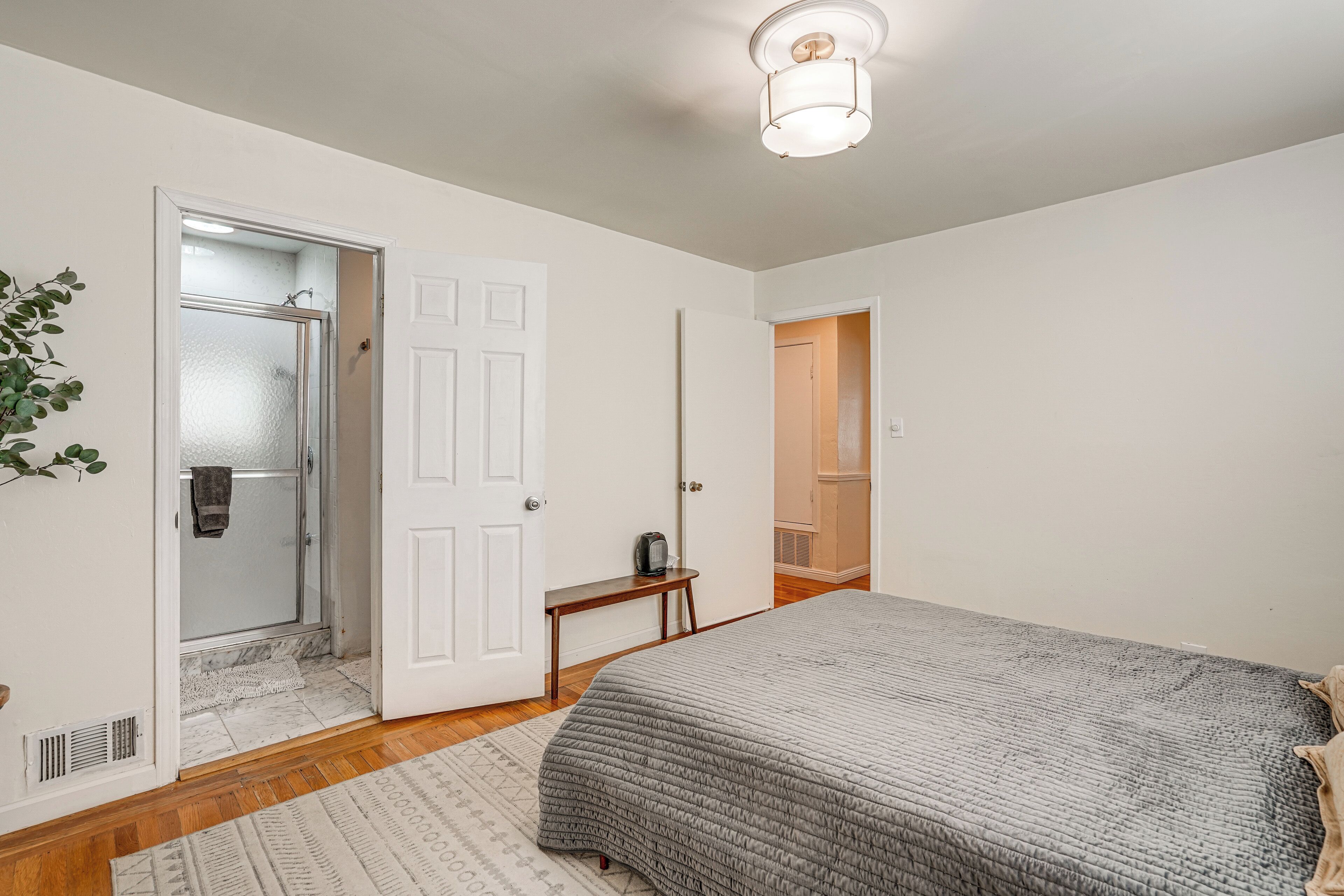 A bedroom with a bed, a door, a mirror, a small table, and a plant.