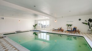 An indoor swimming pool with a tiled floor, a lounge area with chairs, and a door leading outside.