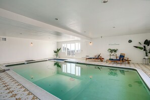Una piscina interior con suelo de baldosas, una zona de descanso con sillones y una puerta que da al exterior.