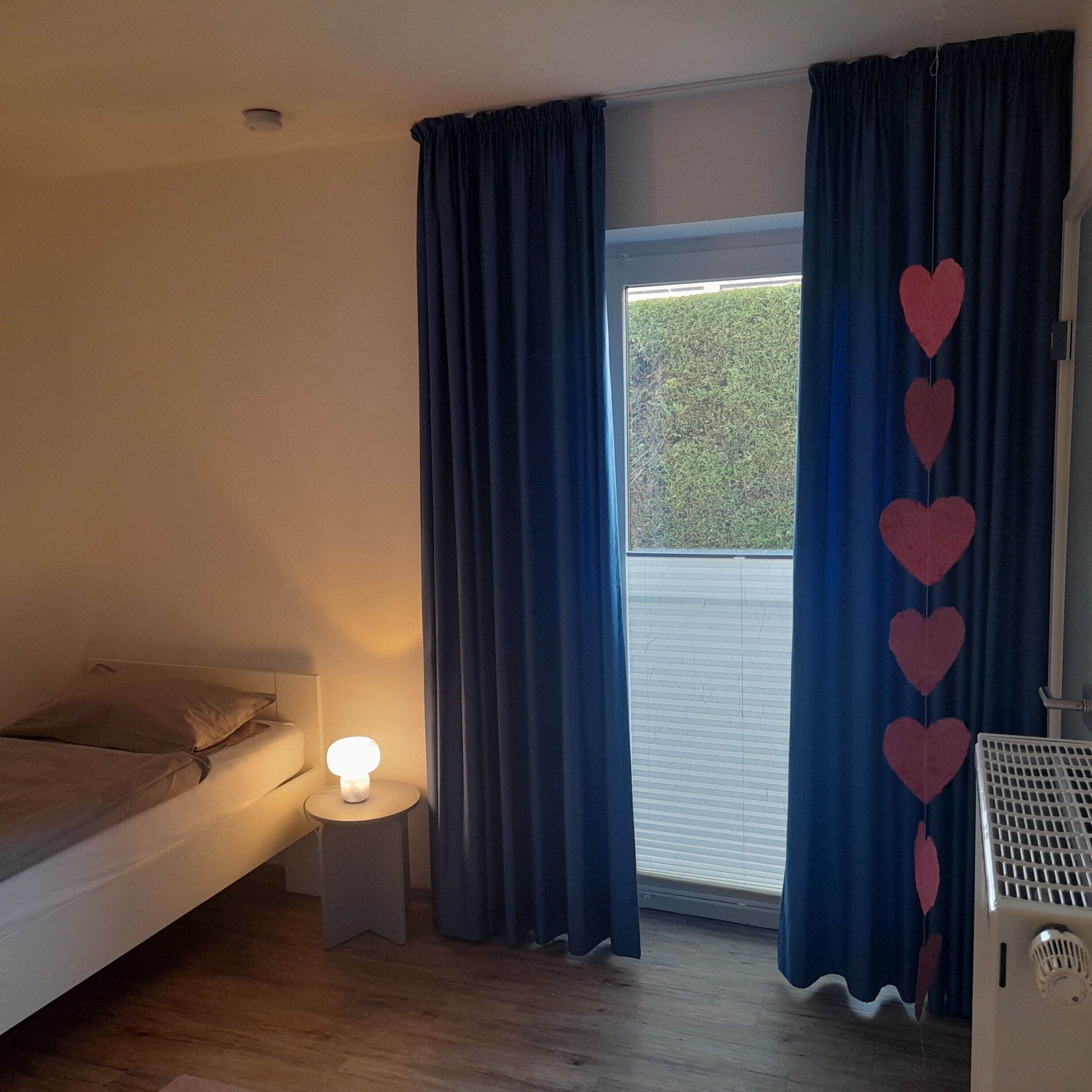 Ein Schlafzimmer mit Bett, Nachttischlampe, einem Fenster mit Vorhängen und einer Heizung.