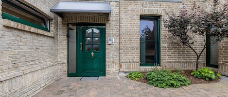 Un edificio de ladrillo con una puerta verde, un pequeño jardín con plantas y una ventana con vistas.