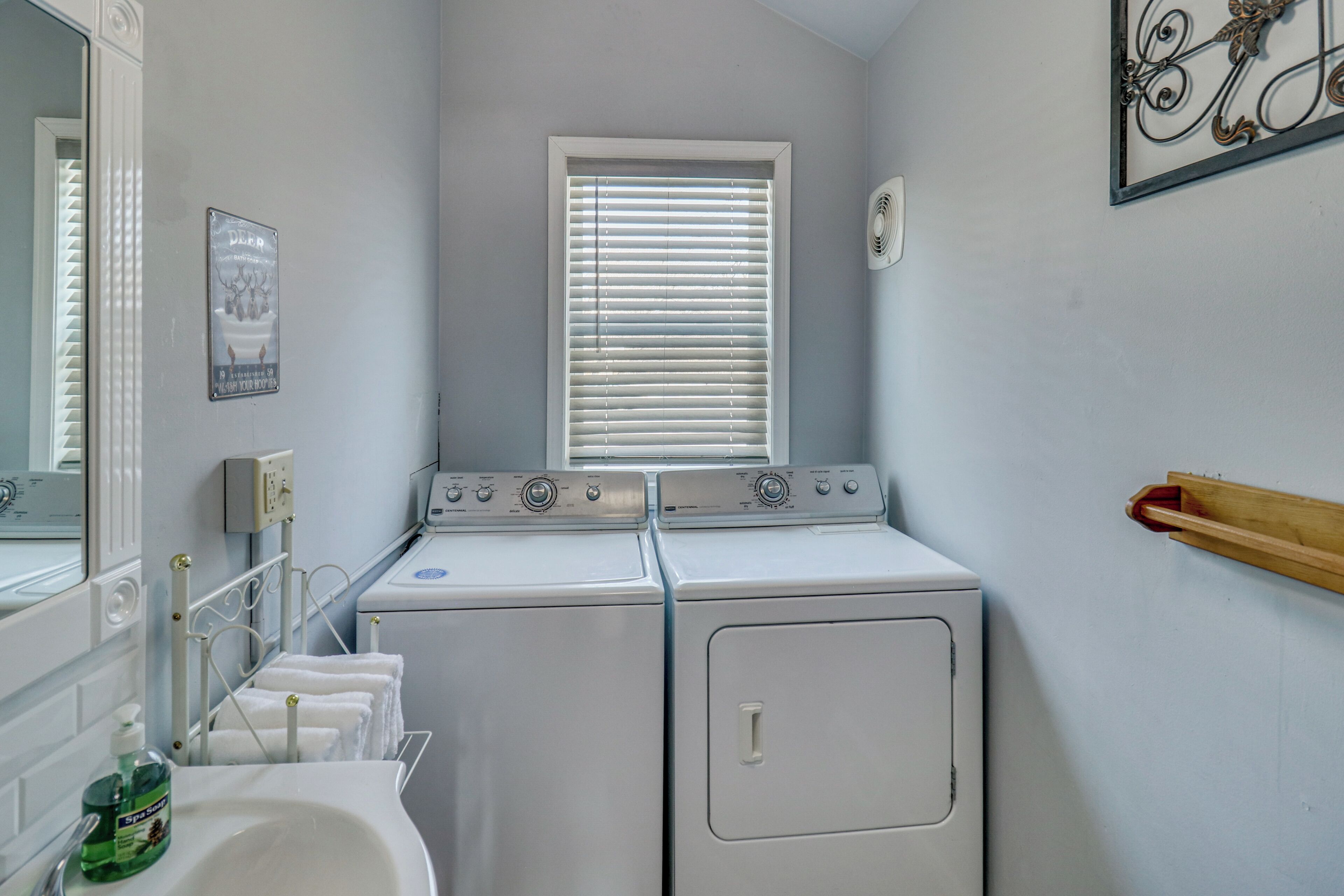 A laundry room with two stacked washers and dryers, a window with blinds, a mirror, and a wall-mounted fan.