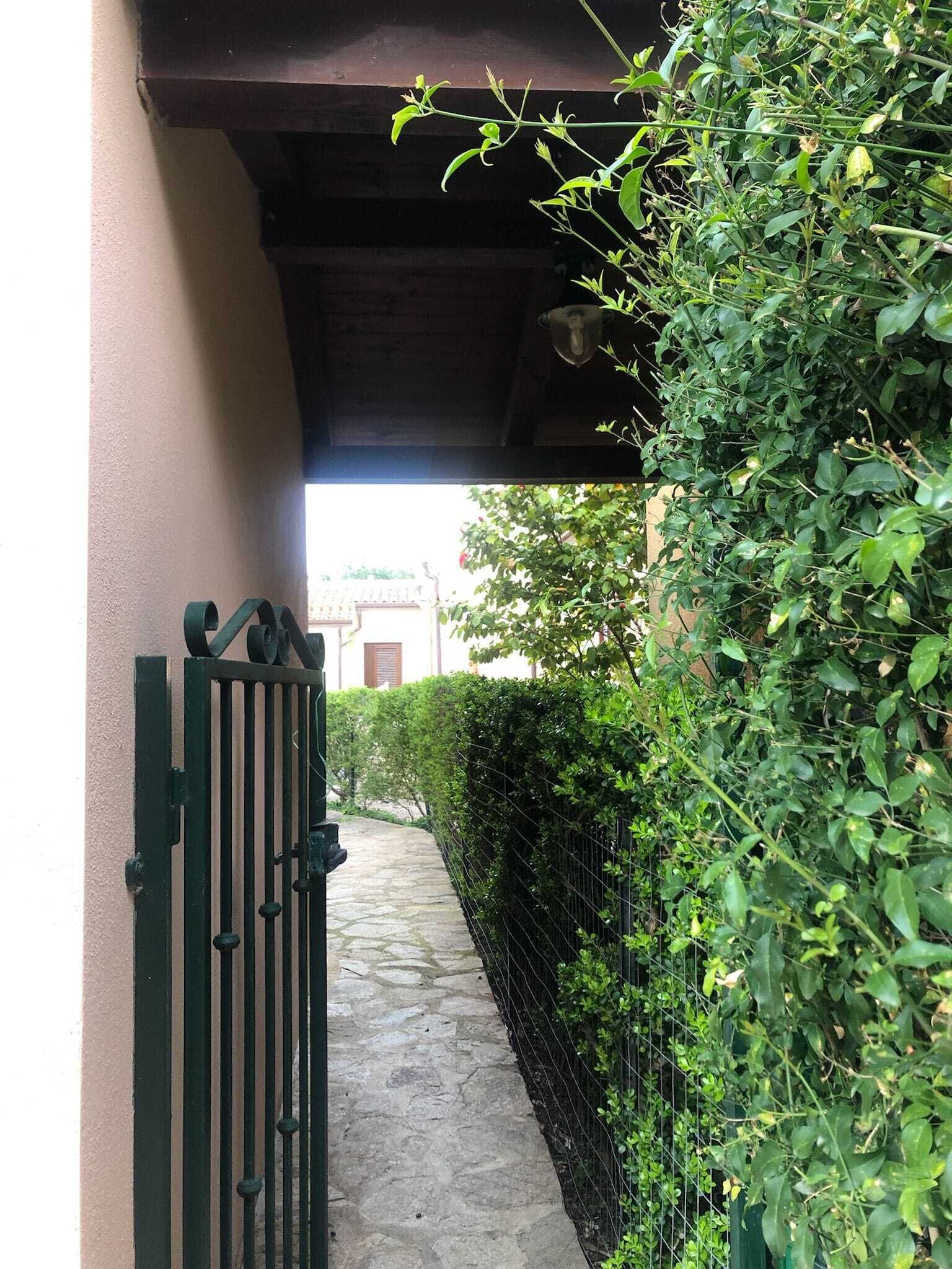 En smal sti der fører til en port, omgivet af frodig vegetation og en bygning med rødt tag i baggrunden.