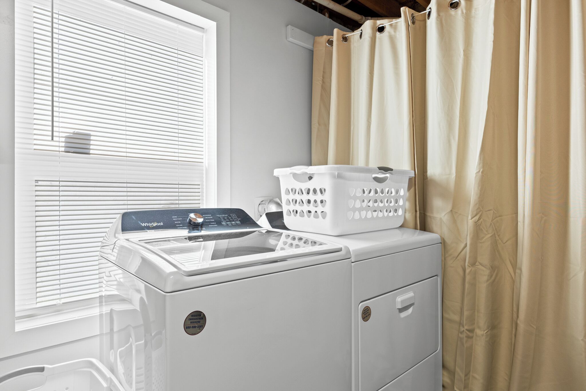 A front-loading washer and dryer unit with a control panel, a white laundry basket, and a beige curtain.