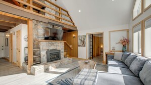 A spacious living room with a stone fireplace, a large gray sofa, and a wooden staircase. - 5 Mi to Soda Springs Mtn Resort: Forest-view Gem! (Truckee)