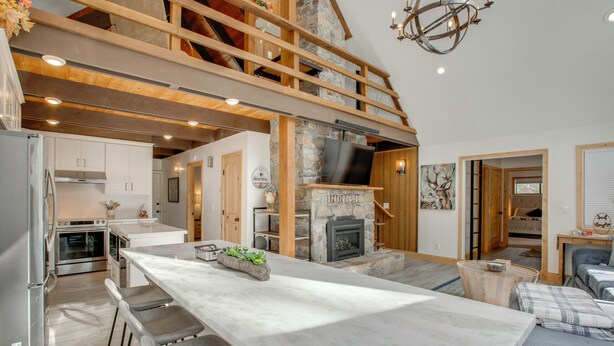 A spacious kitchen with a large island, a stone fireplace, and a loft area with wooden beams. - 5 Mi to Soda Springs Mtn Resort: Forest-view Gem! (Truckee)