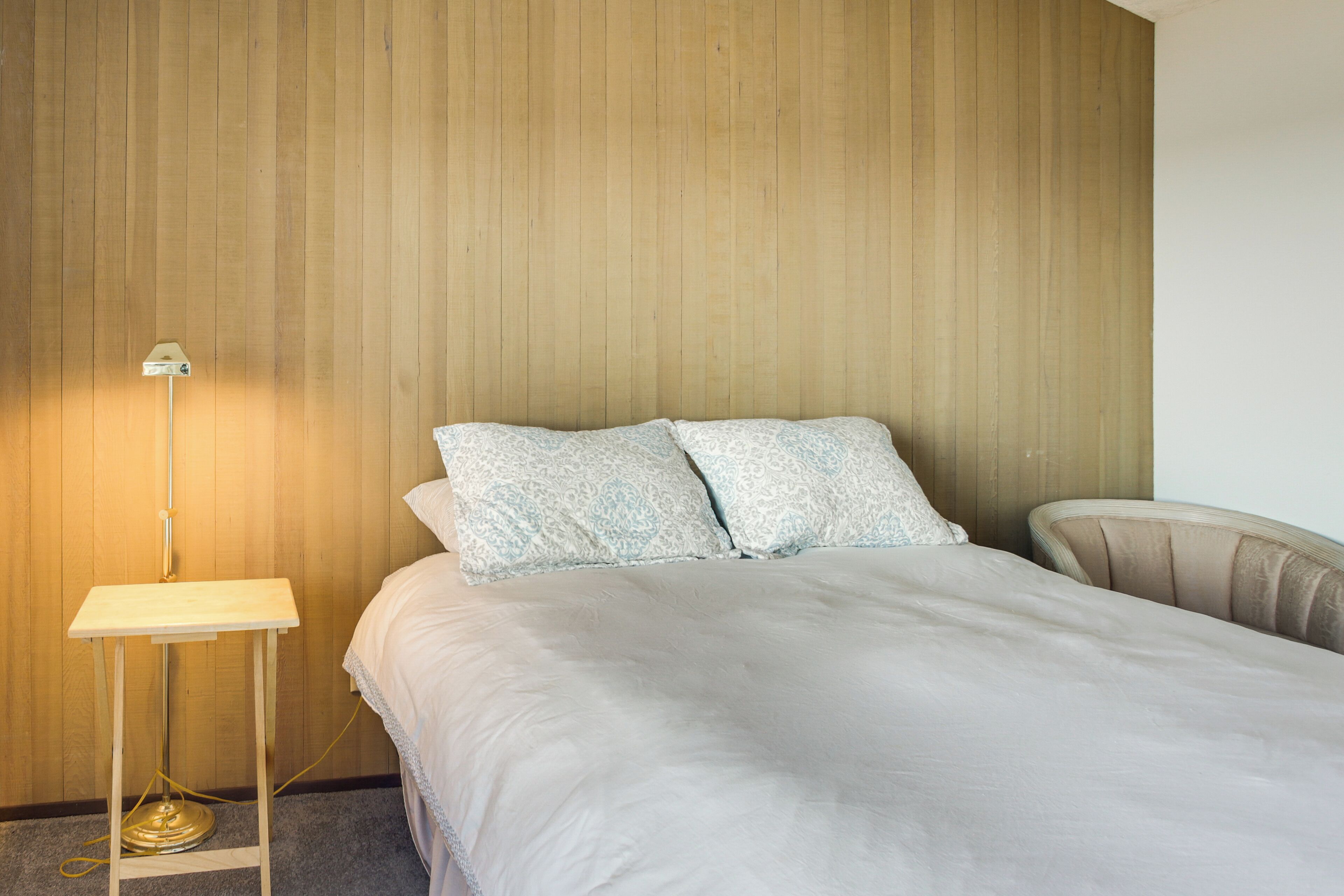 Un dormitorio con una cama, dos almohadas, una mesita de noche con lámpara y una silla.