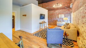 A modern living room with a brick wall, wooden flooring, a blue armchair, a beige sofa, and a black and white patterned rug.