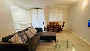 Ein Wohnzimmer mit einem Ecksofa, einem Glastisch, einer Essecke mit Tisch und Stühlen sowie einem an der Wand befestigten Fernseher.
