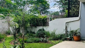 Ein Garten mit einer gefliesten Terrasse, verschiedenen Pflanzen und einer Wand mit einem Fenster.