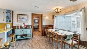 A cozy room with a wooden dining table, chairs, a kitchen area with a counter and stools, and a window with blinds. - Walk to Pigeon Hill Bay: Coastal Maine Cottage (Steuben)