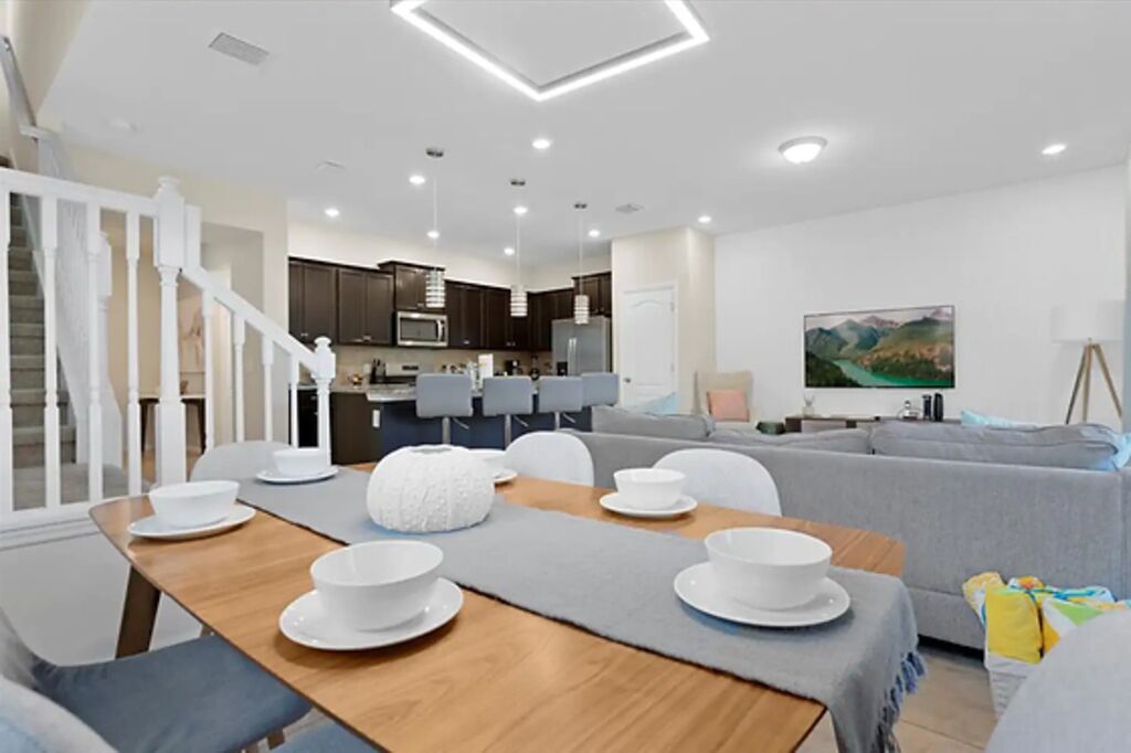 A modern living room with a dining area, a kitchen in the background, and a staircase leading to another level.