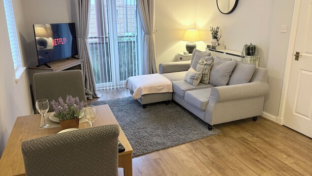 A modern living room with a sofa, a TV, a dining table with a chair, a small side table, and a lamp. - The Kinfirth - cosy beach apartment (Ayr)