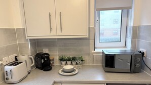 A kitchen with white cabinets, a toaster, a coffee maker, a microwave, and a window with a view. - The Kinfirth - cosy beach apartment (Ayr)