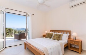 Ein Schlafzimmer mit einem Bett, zwei Nachttischen, einem Deckenventilator und einer Glastür, die auf einen Balkon mit Blick führt.
