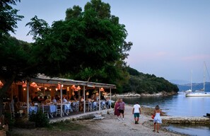 Ein Restaurant im Freien mit Tischen und Stühlen, das auf ein Gewässer und eine hügelige Landschaft blickt.