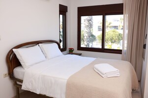 Una cama bien hecha con sábanas blancas y una manta color beige, una pequeña planta en maceta sobre la mesita de noche y una ventana con cortinas.