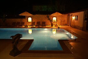 Una piscina con sillas de descanso y sombrillas, rodeada de muros de piedra y un edificio con puertas y ventanas.