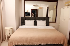 Una cama bien hecha con cabecera, una mesita de noche con cajones y un espejo grande sobre la cama.