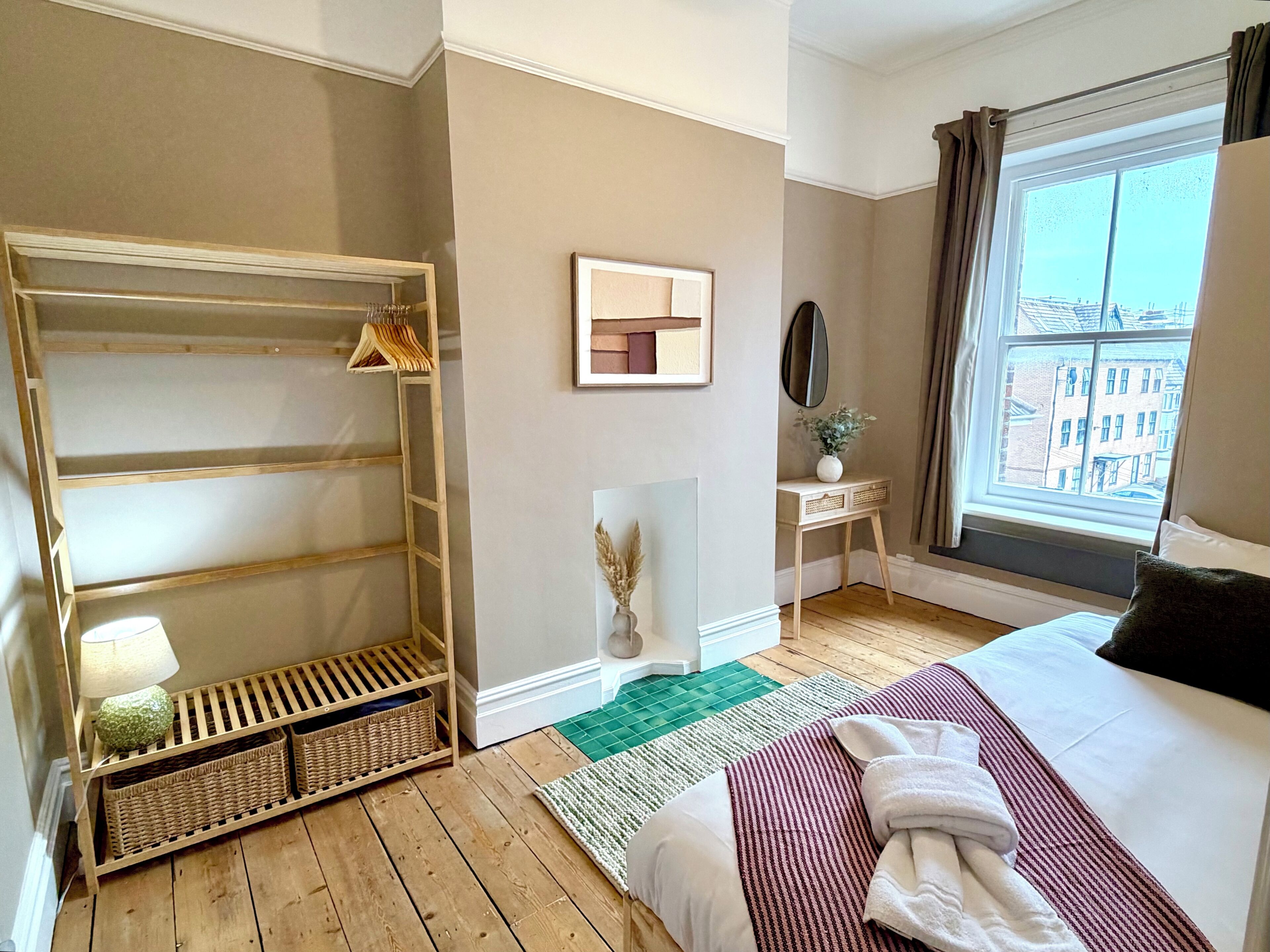 Una habitación de hotel con piso de madera, una cama con manta a rayas, una mesita de noche con un florero, una ventana con cortinas y un espejo montado en la pared.