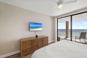 A bedroom with a large bed, a television on the wall, a wicker dresser, and a balcony with a view of the ocean. - Bluewater 1503 by Liquid Life (Orange Beach)