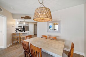 A dining area with a large wooden table, wooden chairs, a hanging light fixture, a kitchen in the background, and a framed mirror on the wall. - Bluewater 1503 by Liquid Life (Orange Beach)