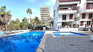 A swimming pool with a metal ladder, surrounded by a tiled patio, palm trees, and a multi-story building with balconies. - Librada Home Playa Las Americas (Playa de las Américas)