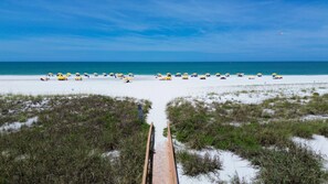 Ein Strand mit einer Holzpromenade, die zum Wasser führt, umgeben von Dünen und gelben Sonnenschirmen.