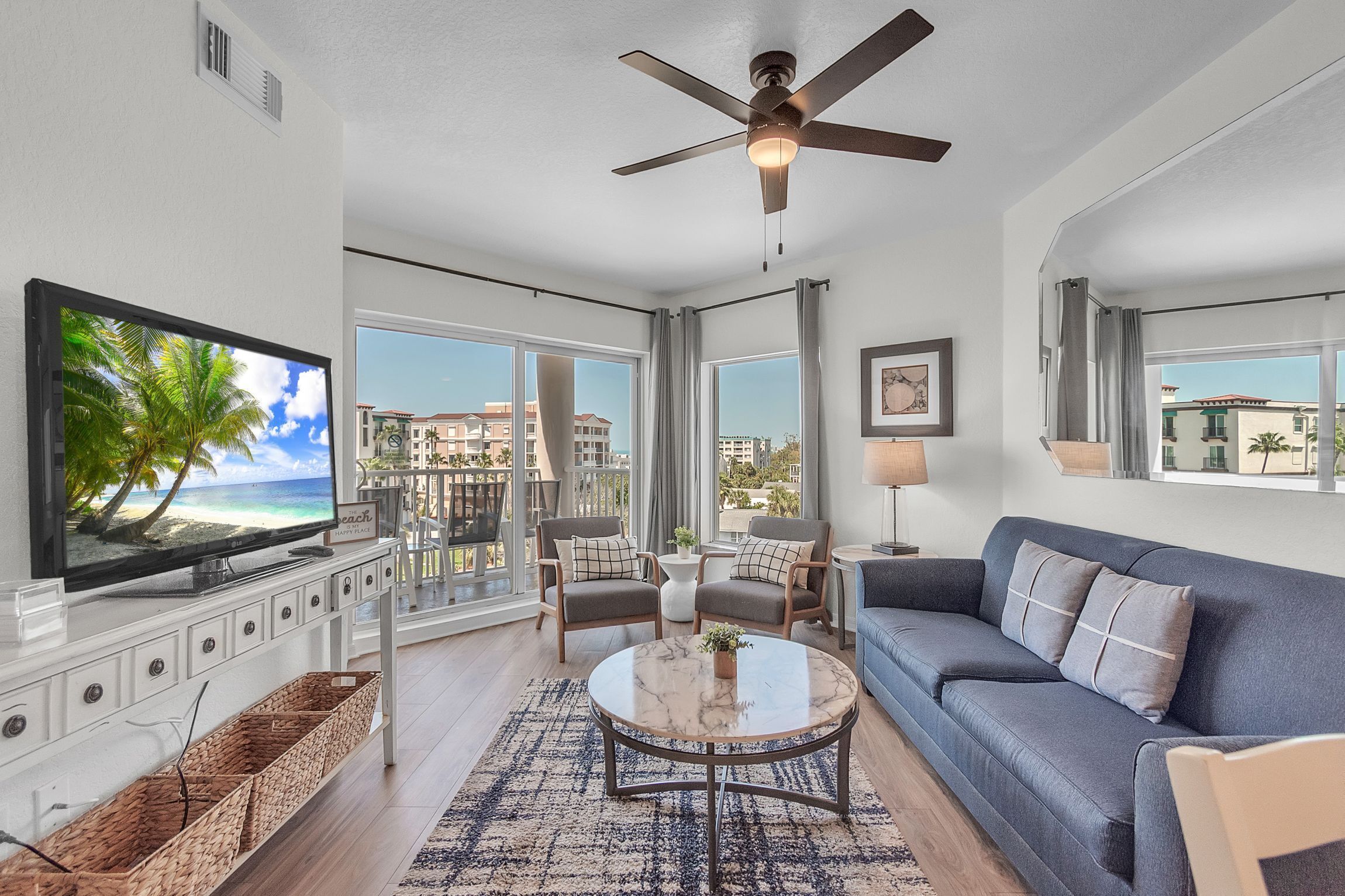 A spacious living room with a large flat-screen TV, a ceiling fan, and a balcony with a view of the city.