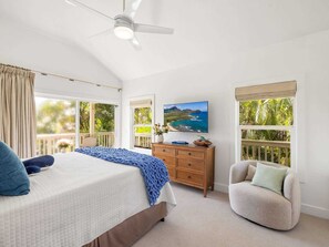 A bedroom with a bed, a chair, a dresser, a ceiling fan, and a large window with a view of a beach. - Poipu Kai - 6BR Villas, Pool - Sleeps 16 (Koloa)