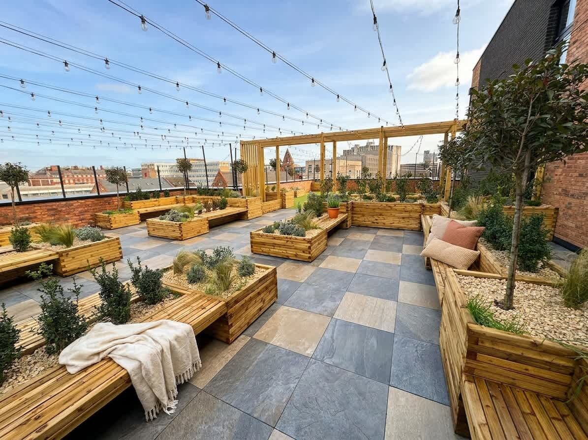 Zona d'estar a l'exterior amb jardineres de fusta, bancs, una estructura de pèrgola i vista a la ciutat.