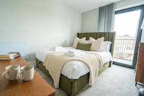 A hotel room with a bed, a table with two mugs, a window with a view, and a door.