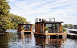 Two modern floating houses with glass windows, wooden decks, and potted plants. - Maréa Stays 1 |Habitation Flottante |Yacht Club TR (Trois-Rivières)