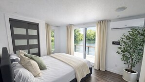 Une chambre avec un grand lit, une porte coulissante, une plante en pot et une vue sur une étendue d’eau.