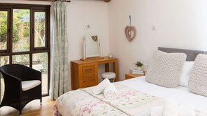 A bedroom with a skylight, wooden beams, a bed with floral bedding, a bedside table, and a chair. - Buttercup - Super King Ensuite Room in Sidlesham (Chichester)