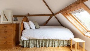 A bedroom with a sloped ceiling, a bed with white bedding, a wooden dresser, a small table, and a window. - Wren Barn - Cosy 2-bed Converted Barn in Sidlesham (Chichester)
