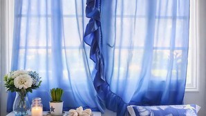 A bedroom with a white bed, a white nightstand with a vase of flowers, a small plant, a candle, and a decorative pillow. - Kabara Garden Oasis in Aegina BBQ Priv Parking (Aegina)