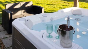 Ett bubbelbad med en metallhink och två glas, placerat under en träpergola med ljusslingor, i en trädgård med en gräsmatta och ett hus i bakgrunden.