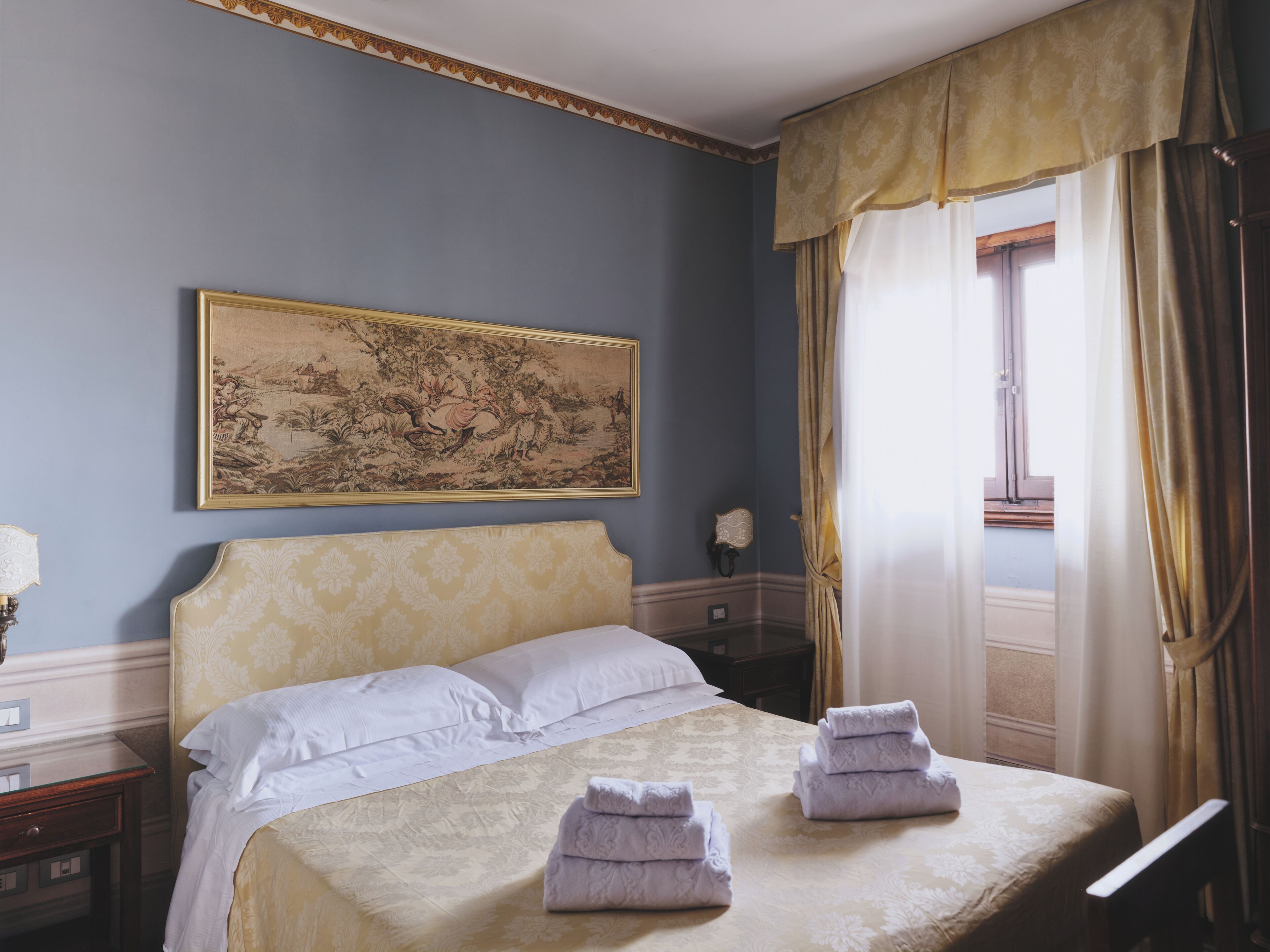 A bedroom with a bed, a nightstand, a lamp, a painting, and a window with curtains.