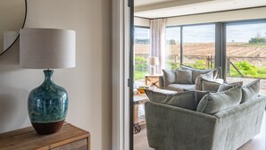 Un saló modern amb un sofà i una làmpada sobre una taula auxiliar de fusta, grans finestres amb vista a un camp, i un paviment de fusta.