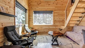 A wooden cabin interior with a sofa, two chairs, a table, and a window with a snowy forest view. - Nature & SPA | Chalet du Sommet (Sainte-Rose-du-Nord)