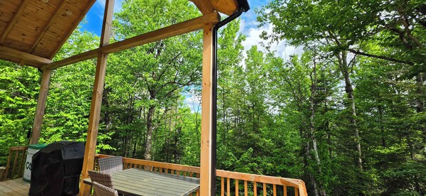 A wooden deck with a grill and wicker furniture, surrounded by dense green forest. - Nature & SPA | Chalet du Sommet (Sainte-Rose-du-Nord)