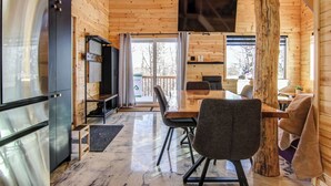A wooden cabin interior with a dining area, a kitchen, and a large window. - Nature & SPA | Chalet du Sommet (Sainte-Rose-du-Nord)