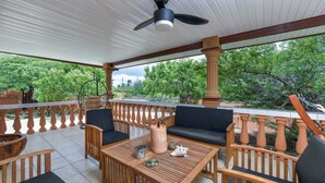 A covered patio with wooden furniture, a ceiling fan, and a view of greenery.