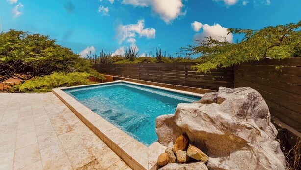 A rectangular swimming pool with clear blue water, surrounded by a stone patio and a wooden fence.