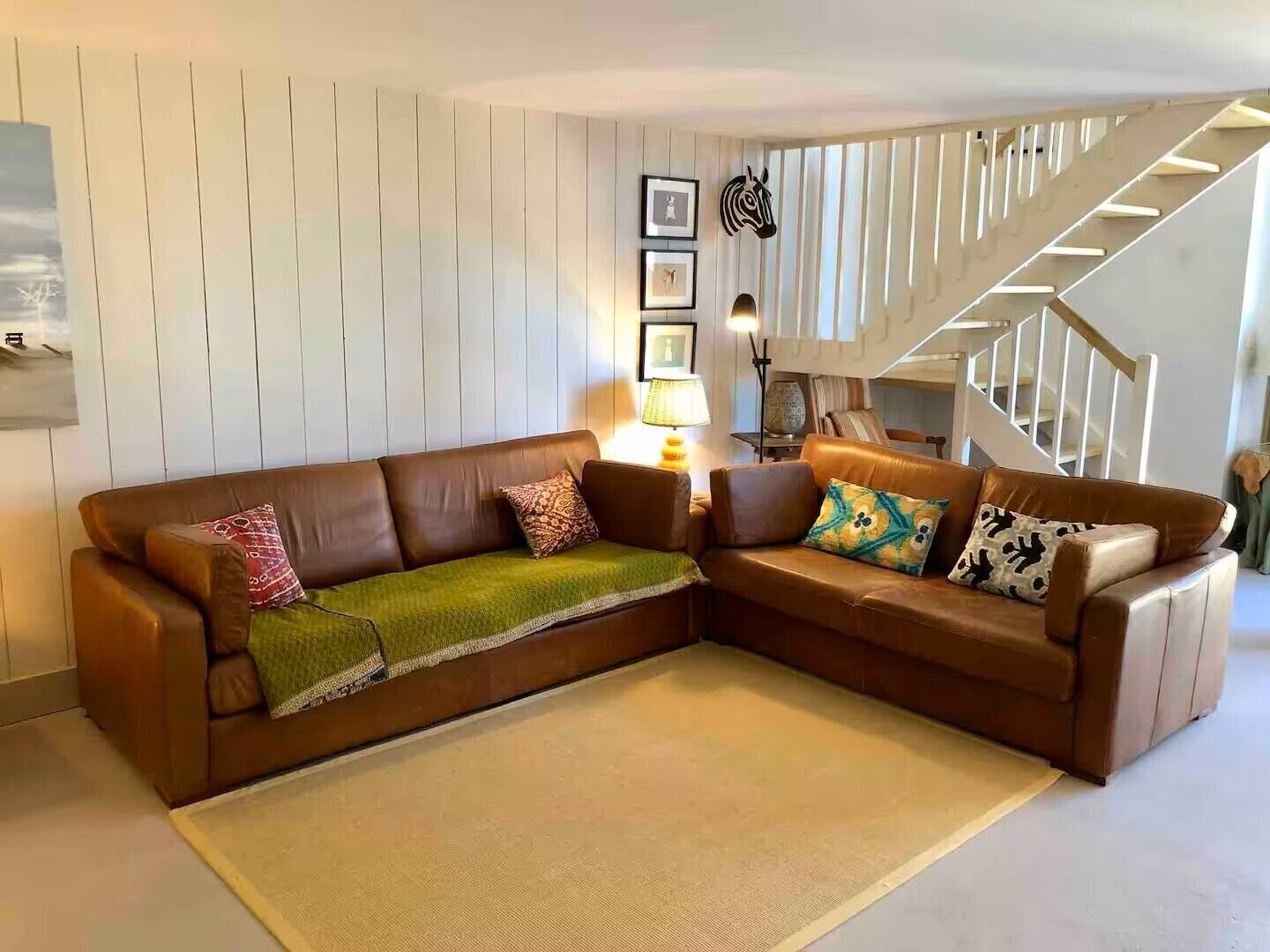 Un salon avec un canapé d’angle en cuir marron, un petit tapis, une lampe et un escalier.