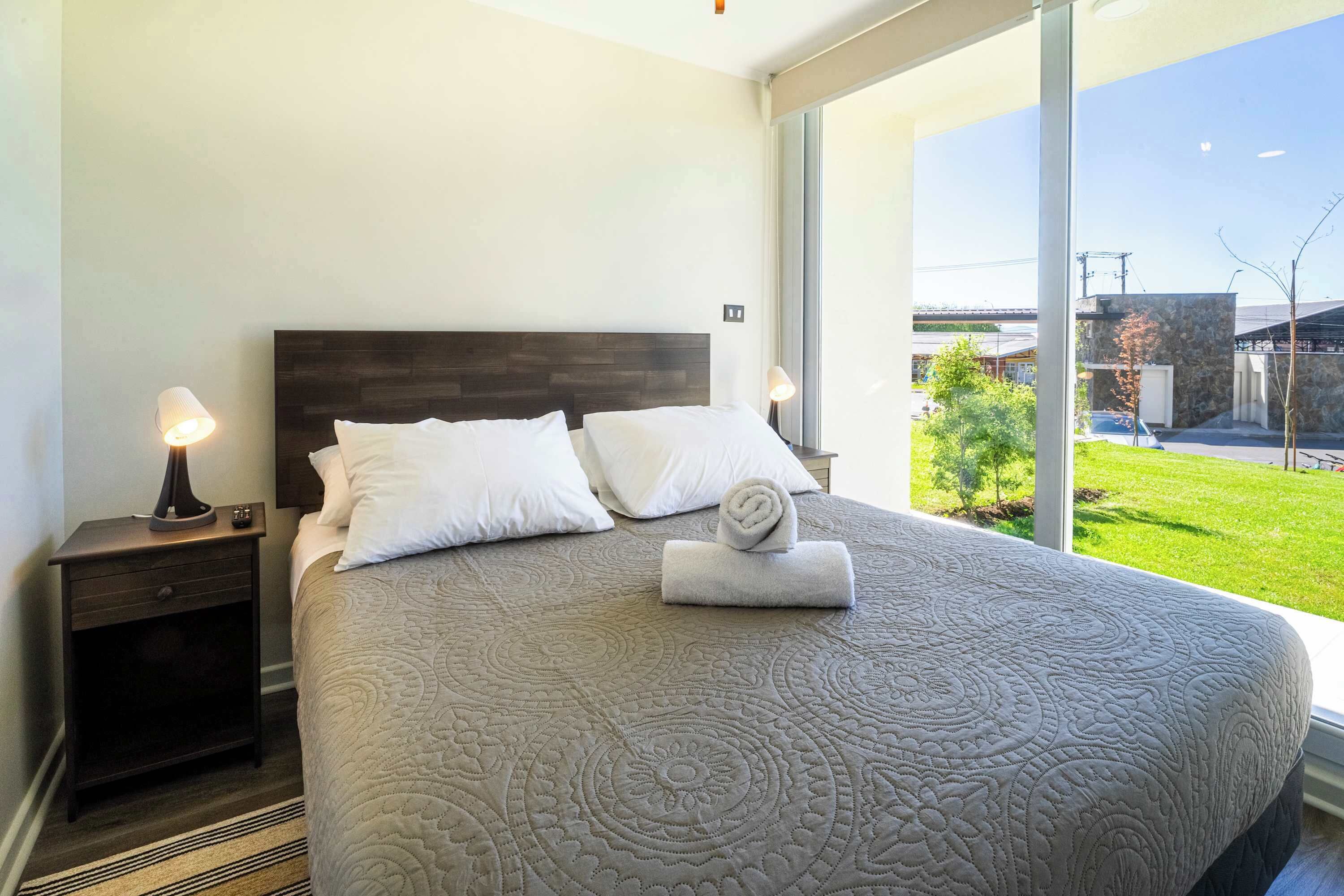 A bedroom with a large bed, two bedside tables, and a view of a green landscape through large windows.