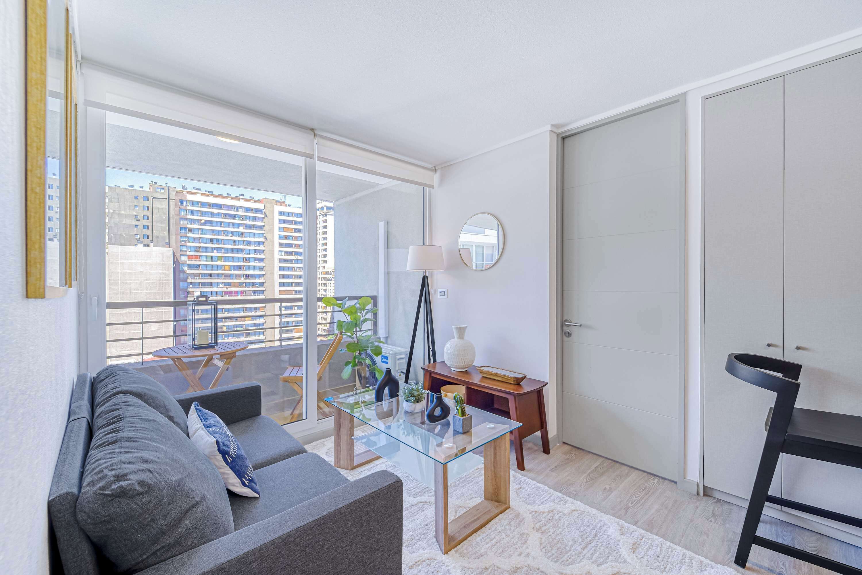 A modern living room with a glass coffee table, grey sofa, and a view of the cityscape through large windows.
