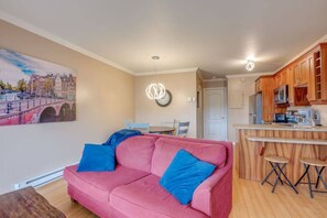 A living room with a pink sofa, a dining area with a table and chairs, and a kitchen with wooden cabinets and a bar area.